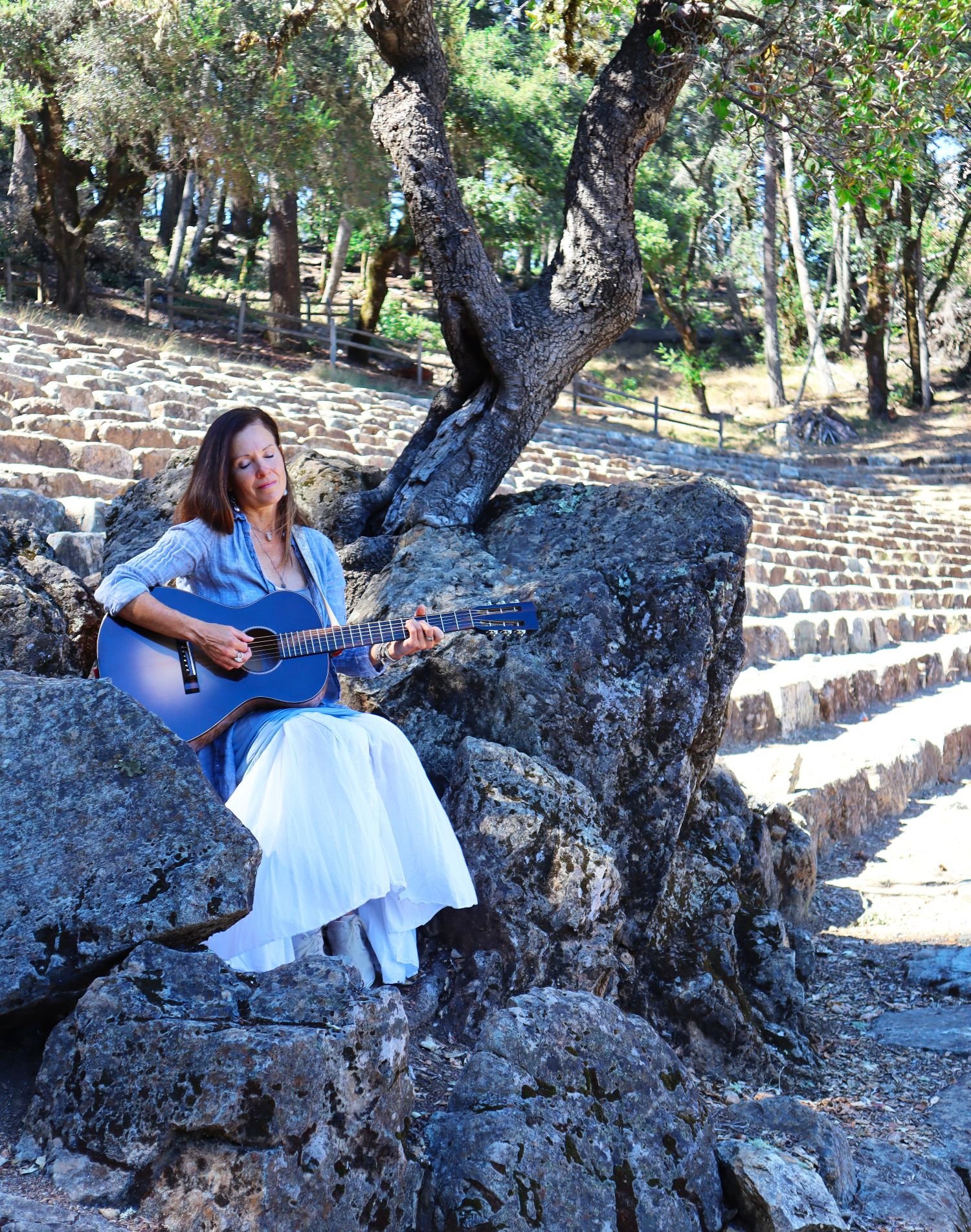 A woman is sitting on rocks outdoors, playing an acoustic guitar with her eyes closed and a serene expression. She is dressed in a blue long-sleeve shirt and a white skirt. Behind her, there are trees, a pathway with stairs, and a rustic wooden fence, suggesting a peaceful natural setting.