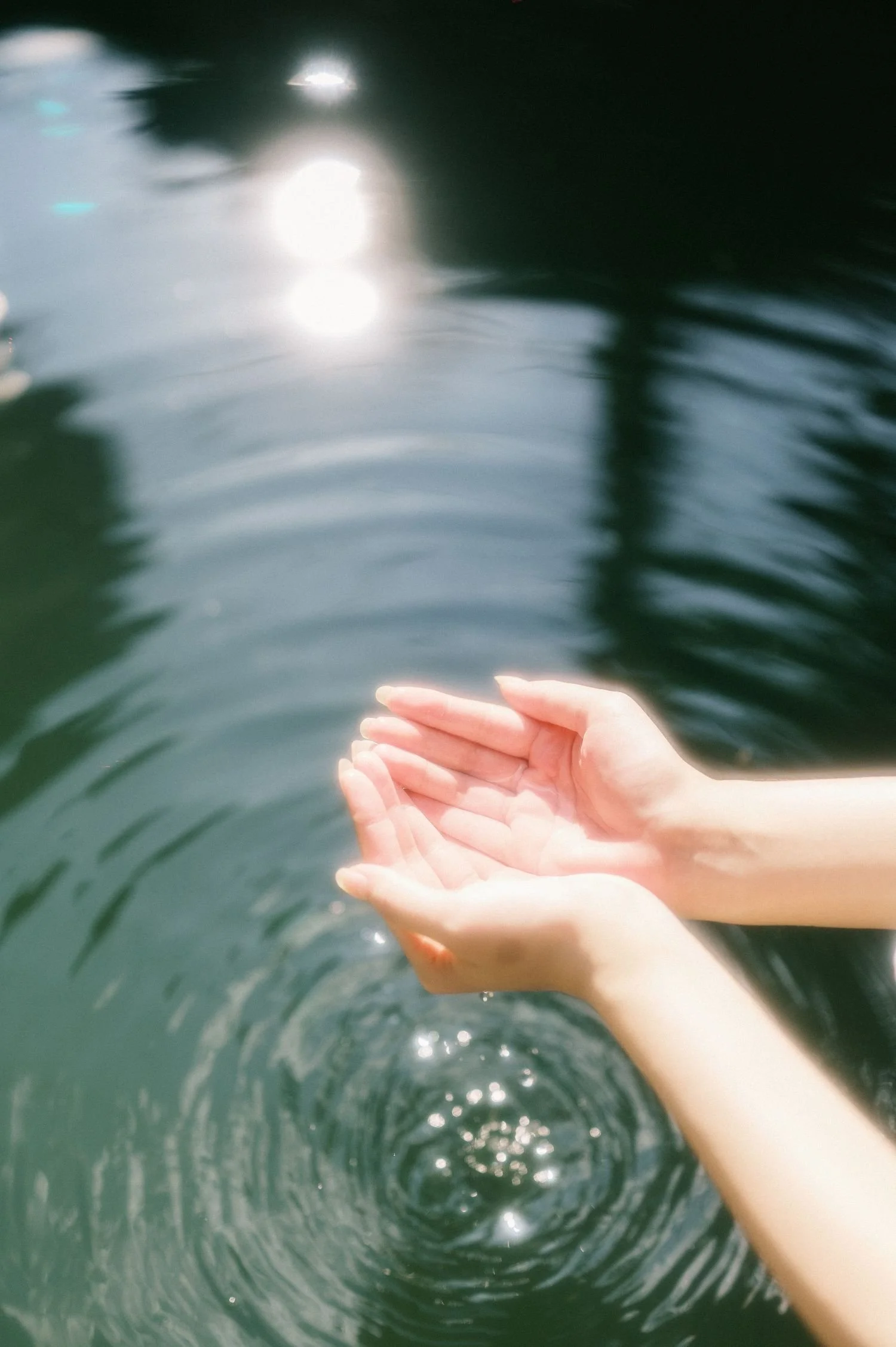 A person's hands are cupped together under water, creating ripples in a body of water with sunlight reflecting on the surface.