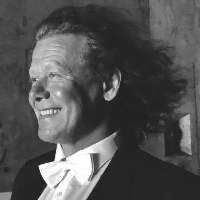 A man with long, wavy hair smiling in black and white, wearing a tuxedo with a bow tie.