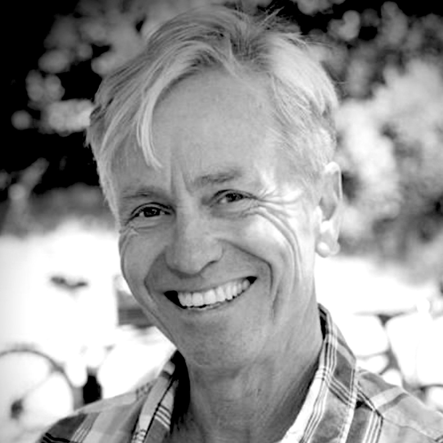 A black and white photo of a smiling woman with short, light-colored hair, wearing a plaid shirt, outside with trees in the background.