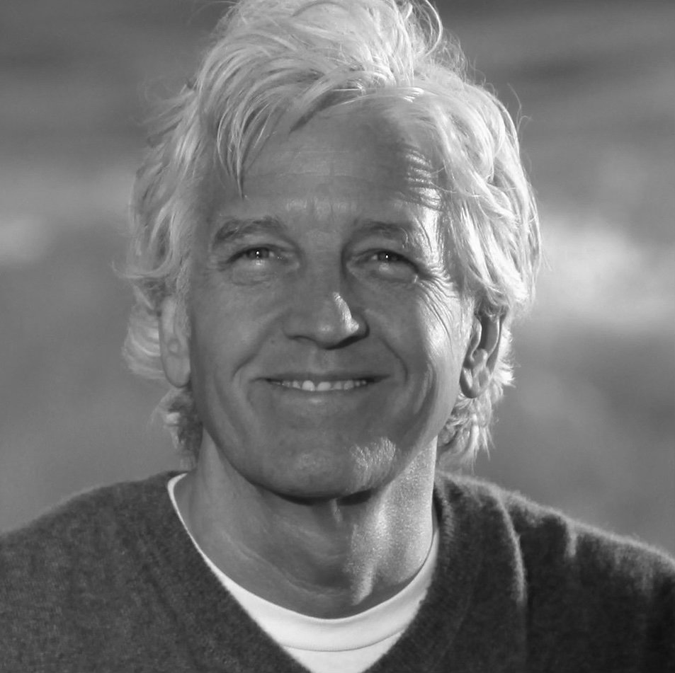 Black and white close-up portrait of a smiling older man with wavy, light-colored hair, wearing a dark sweater over a collared shirt.