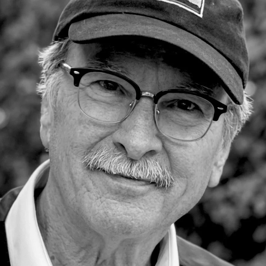 Close-up black-and-white portrait of an older man with glasses, mustache, and a cap, smiling gently.