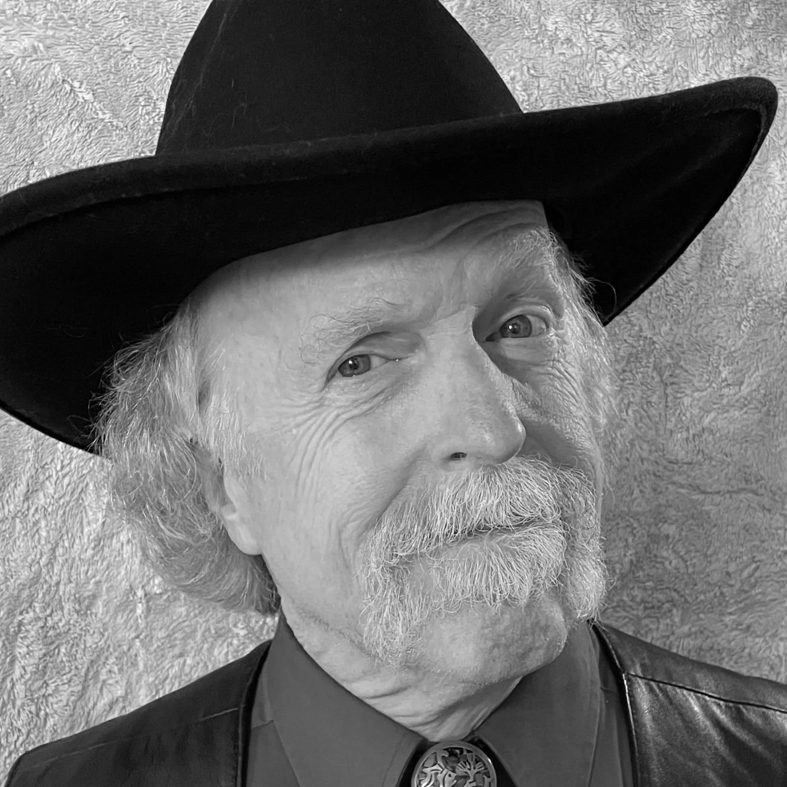 Close-up black-and-white photograph of an elderly man with white hair and beard, wearing a wide-brimmed cowboy hat and a leather jacket, against a textured wall background.