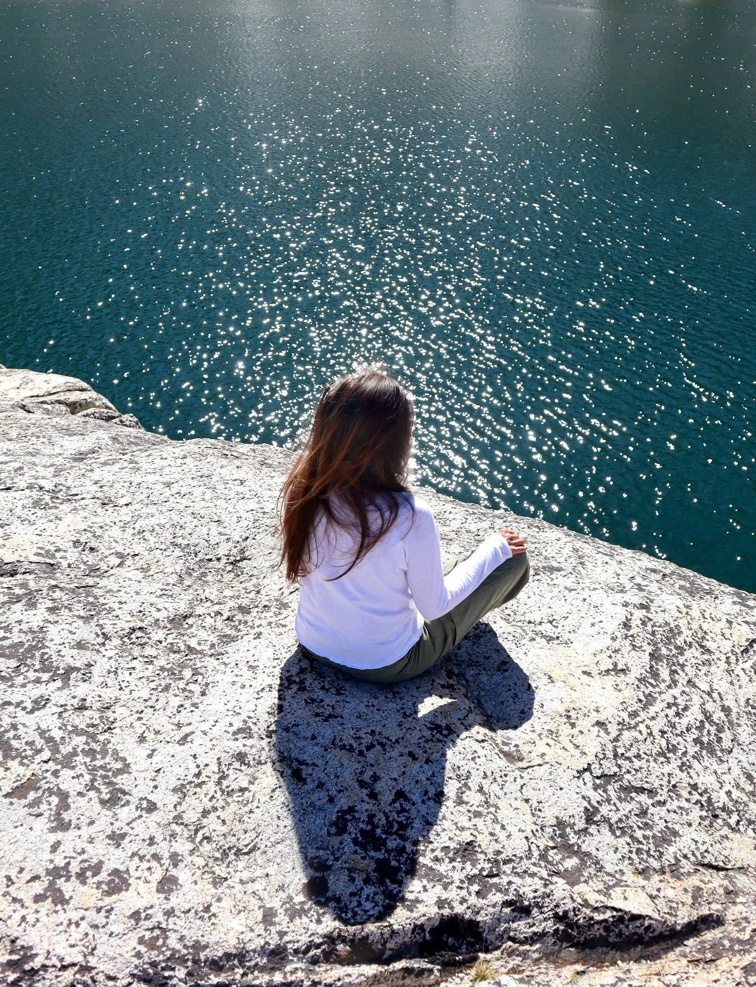 A woman with long brown hair sitting on a rocky ledge beside a large body of water that sparkles in the sunlight.