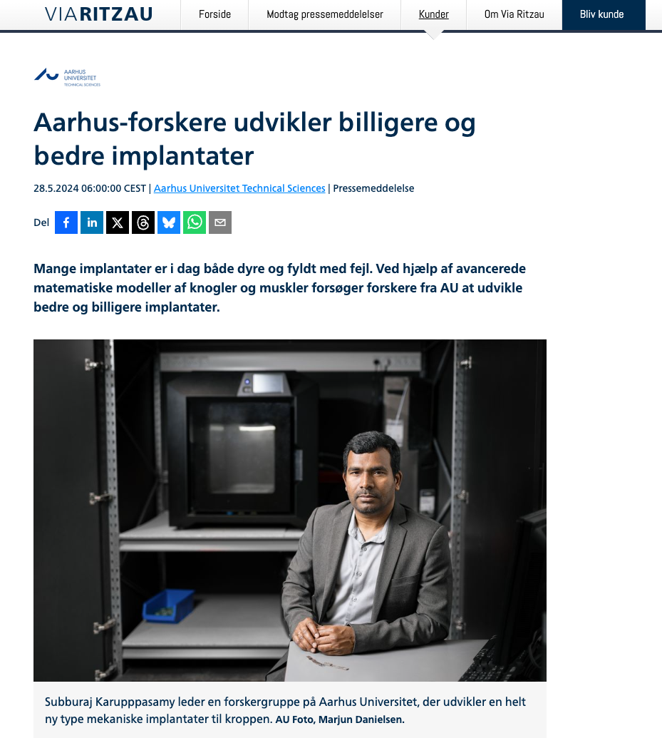 A man in a suit sitting at a desk in front of scientific equipment including a 3D printer and shelves in a laboratory or research environment at Aarhus University.
