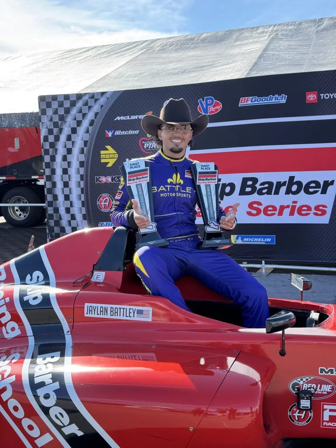 Jaylan Battley smiling with two first-place trophies in his Skip Barber formula car at Road America