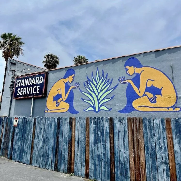 Street mural features two stylized, yellow female figures with blue hair, sitting in a crouched position, facing each other and observing a large plant in the center. A sign on the wall reads "Standard Service," and there is a wooden fence below the mural with a blue paint finish.