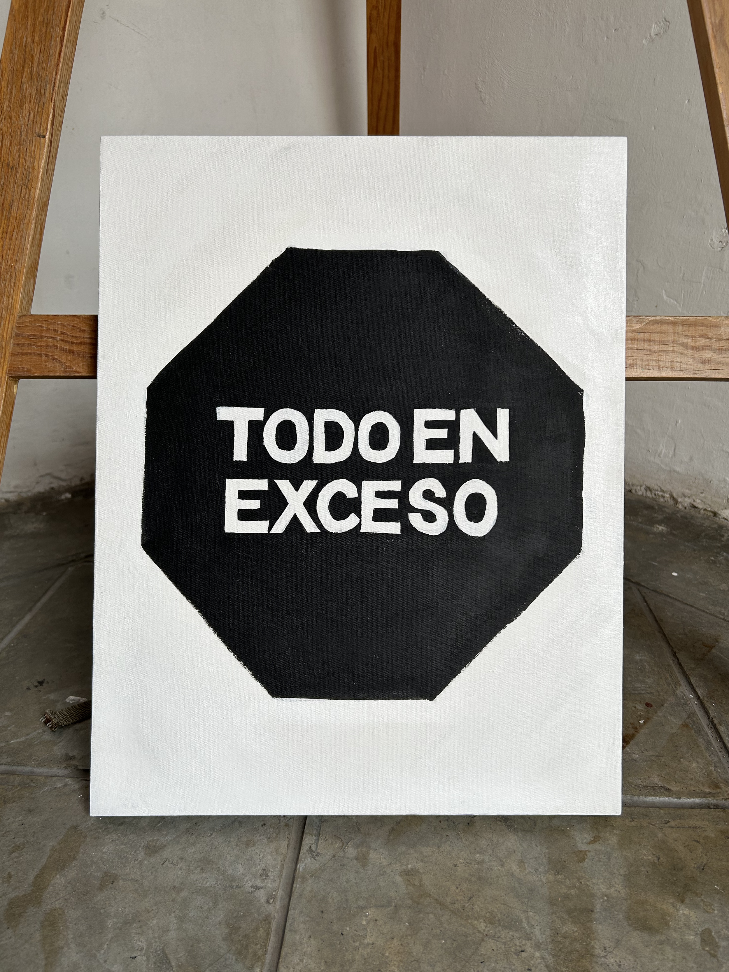 Black and white hand-painted sign with a stop sign shape and Spanish text reading "TODO EN EXCESO" on a wooden chair with a textured wall background.