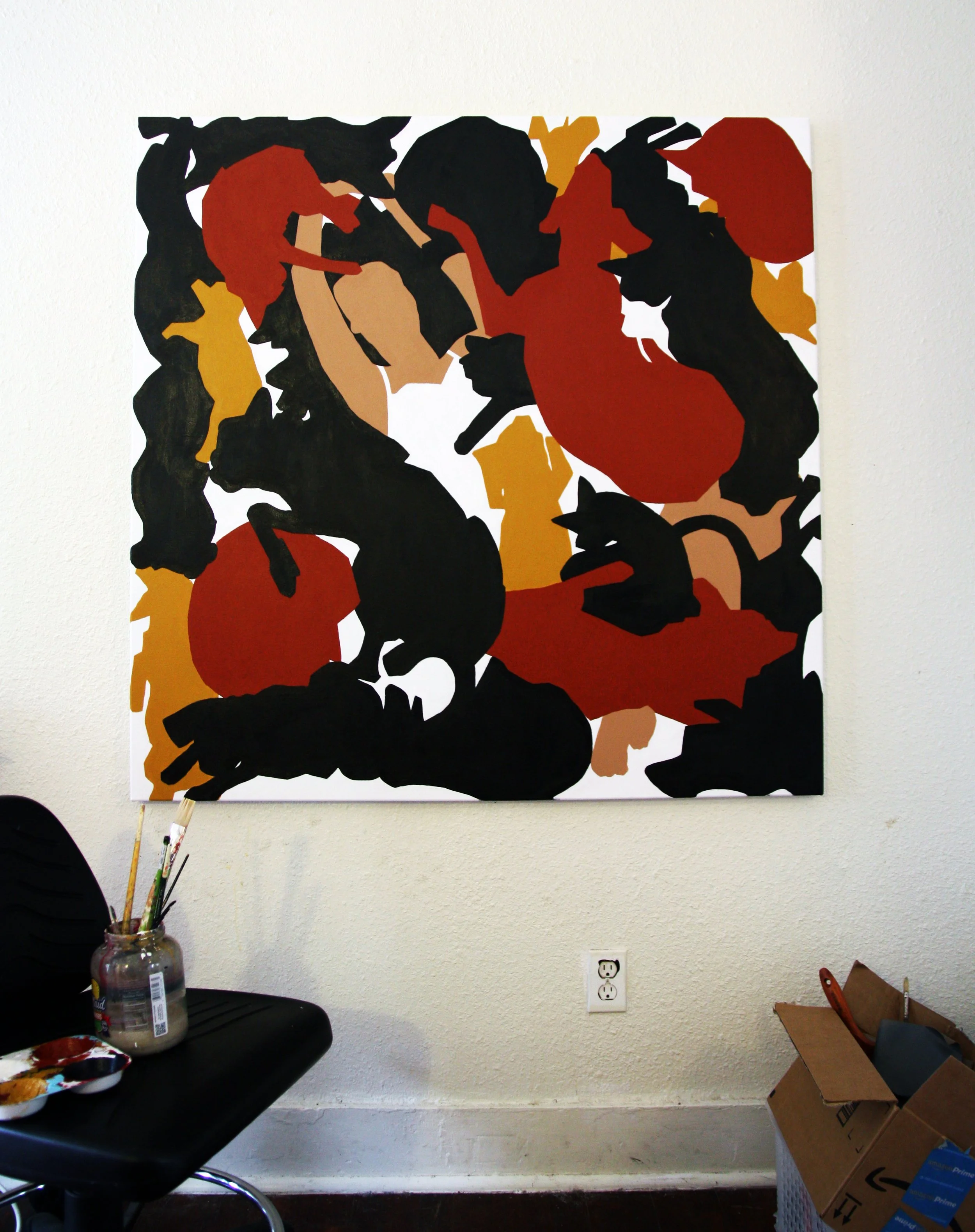 Abstract painting with various black, red, yellow, and beige shapes on a white wall, with art supplies and a cardboard box nearby.