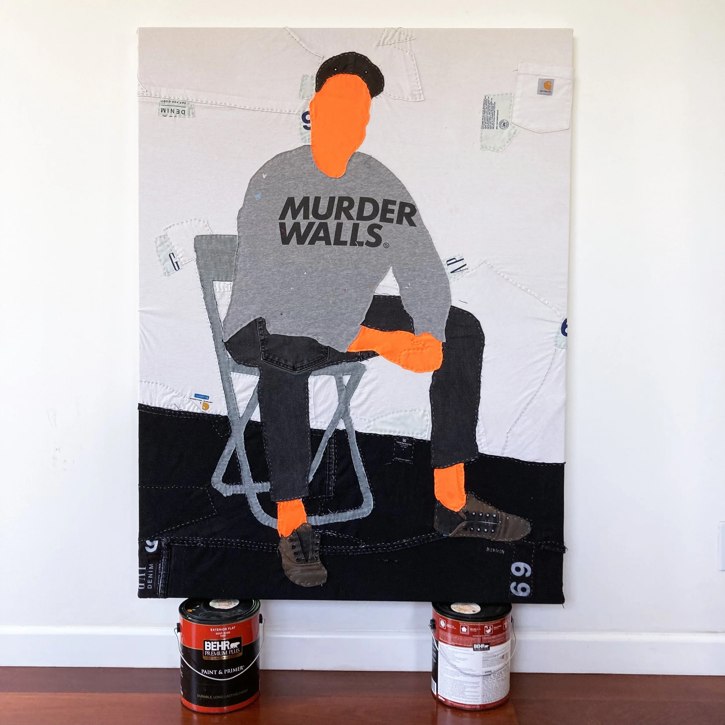 A mixed-media art piece depicting a person with a carrot nose, wearing a gray shirt with the words 'MURDER WALLS' printed on it, sitting on a chair. The art is created with fabric and paint, and has various paint cans at the bottom of the artwork.