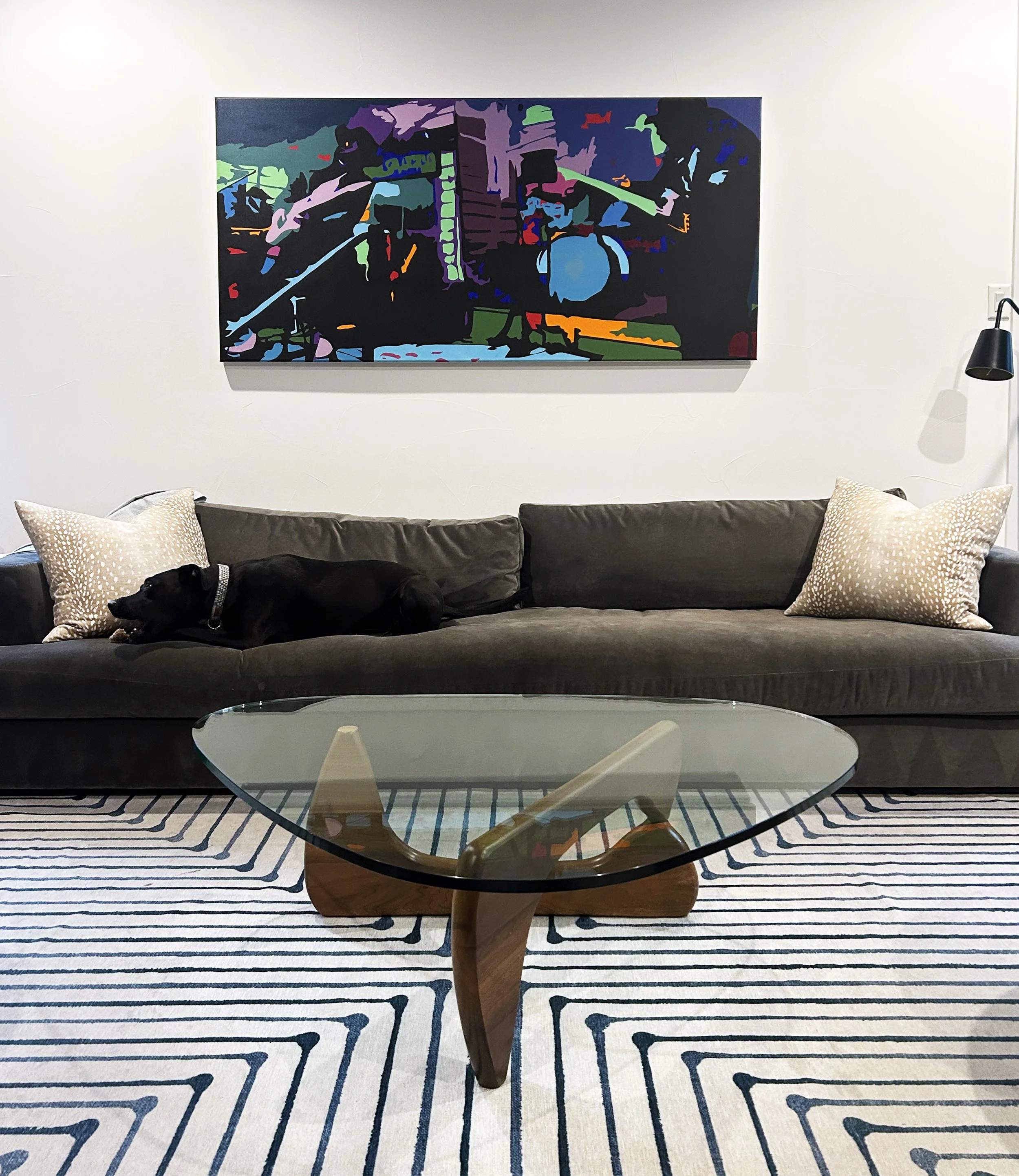 Modern living room with a black sofa, beige patterned pillows, a glass coffee table, a vibrant abstract painting on the wall, a black wall-mounted lamp, and a striped rug.