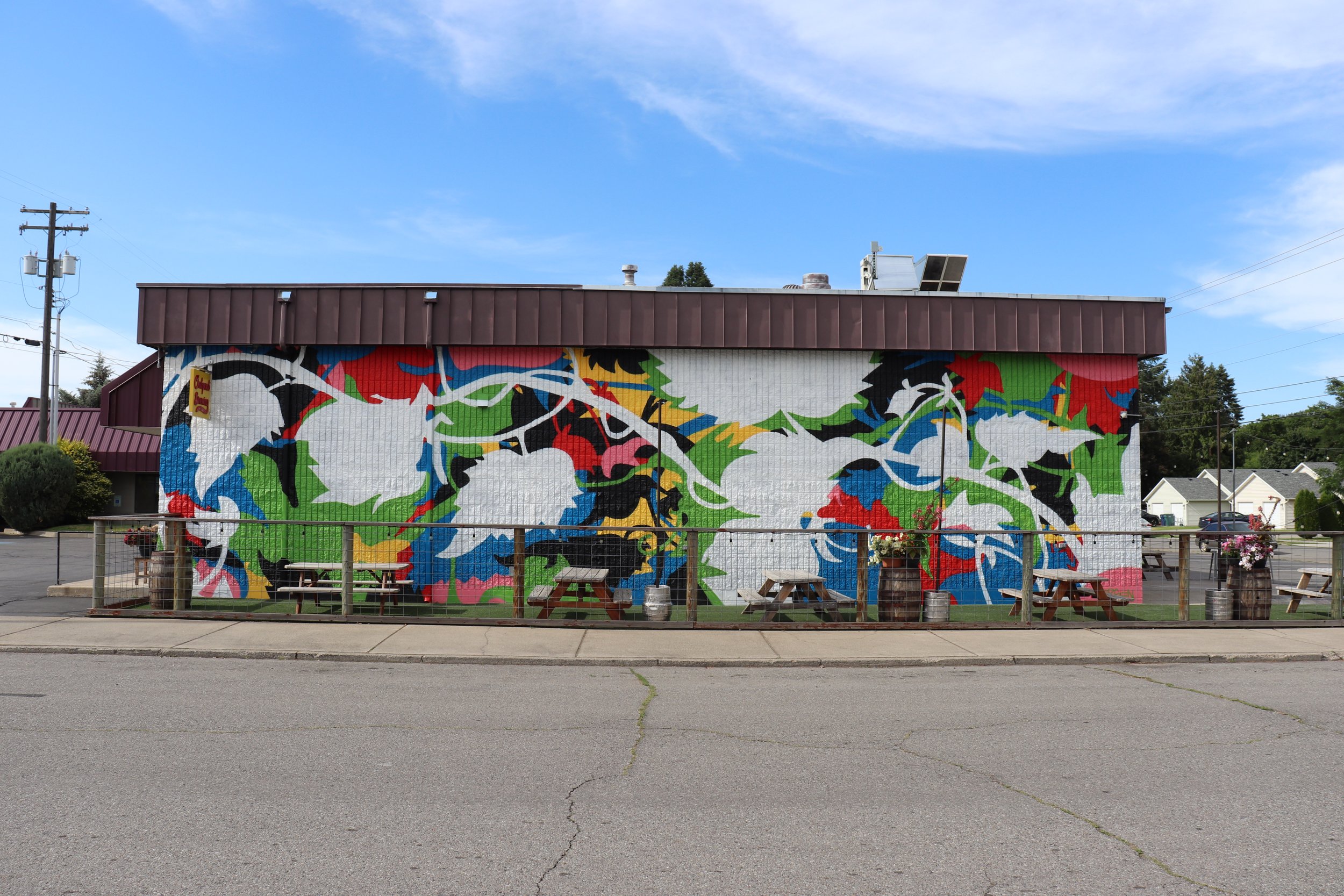 Colorful mural painted on the side of a building, depicting abstract shapes in red, green, blue, black, white, and yellow. Outdoor seating area with picnic tables and barrels in front of the mural, with a blue sky overhead.
