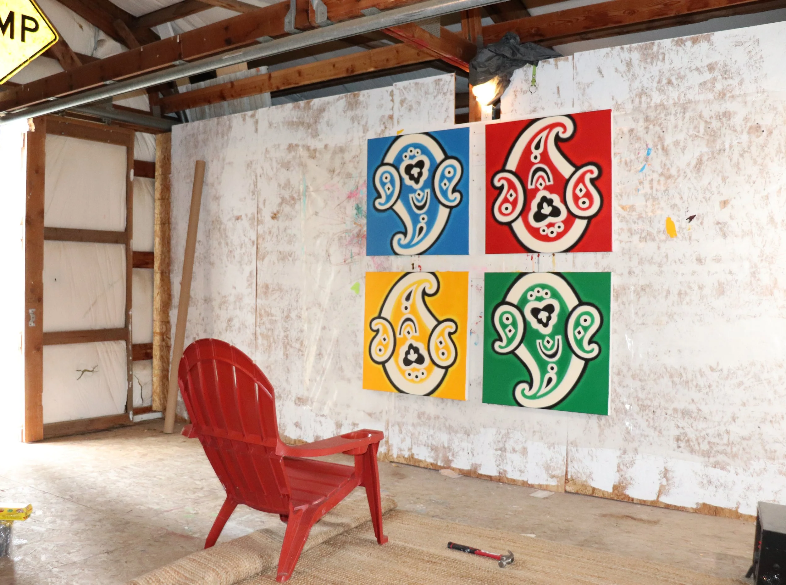Artwork with colorful abstract patterns on a wall in a wooden-paneled room, with a red plastic chair, a sun hat, and a marker on the floor.