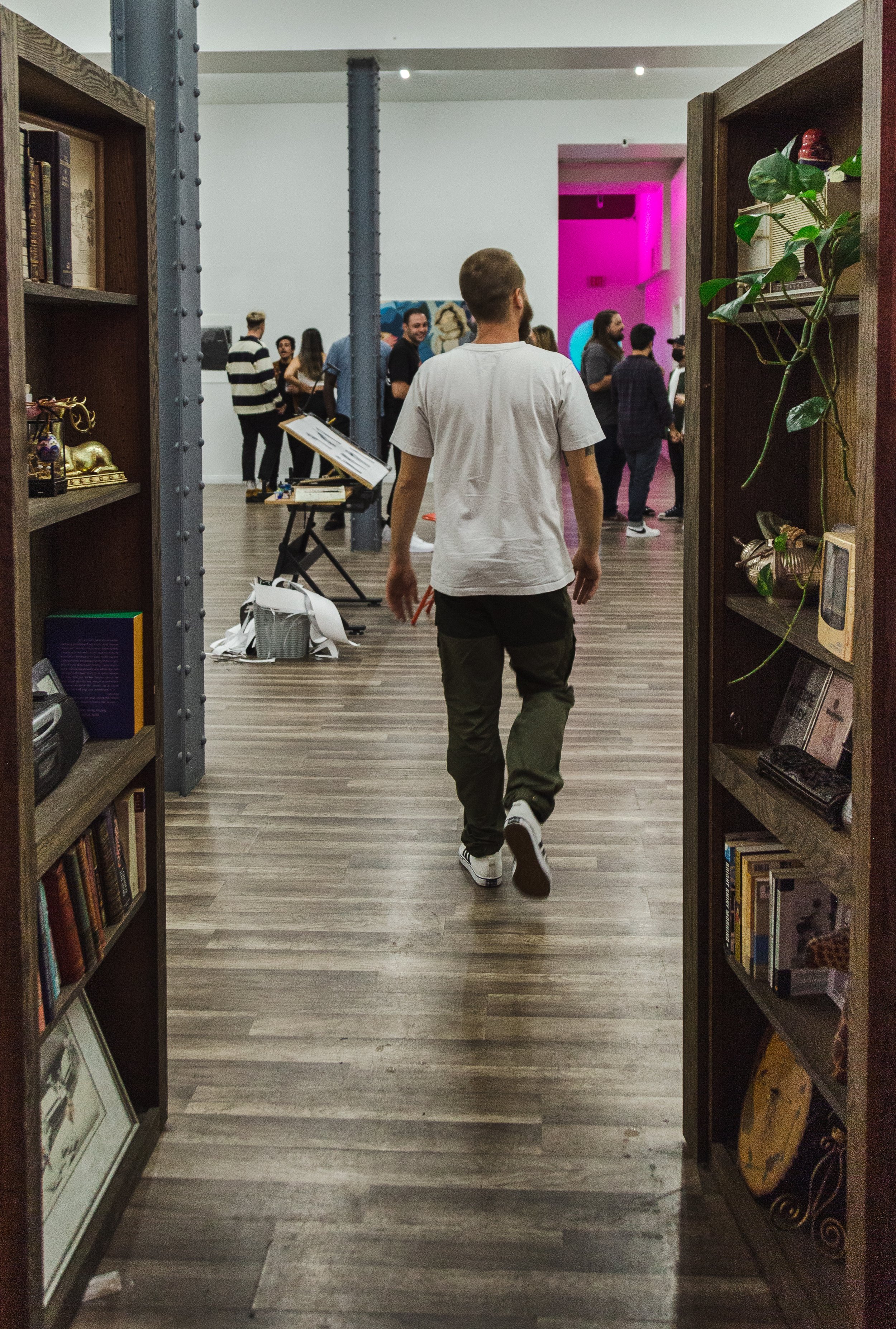 A view through an open doorway into an art gallery or bookstore, with a man walking inside towards a group of people gathered around a display. The space features wooden bookshelves, art, and a pink-lit corridor.