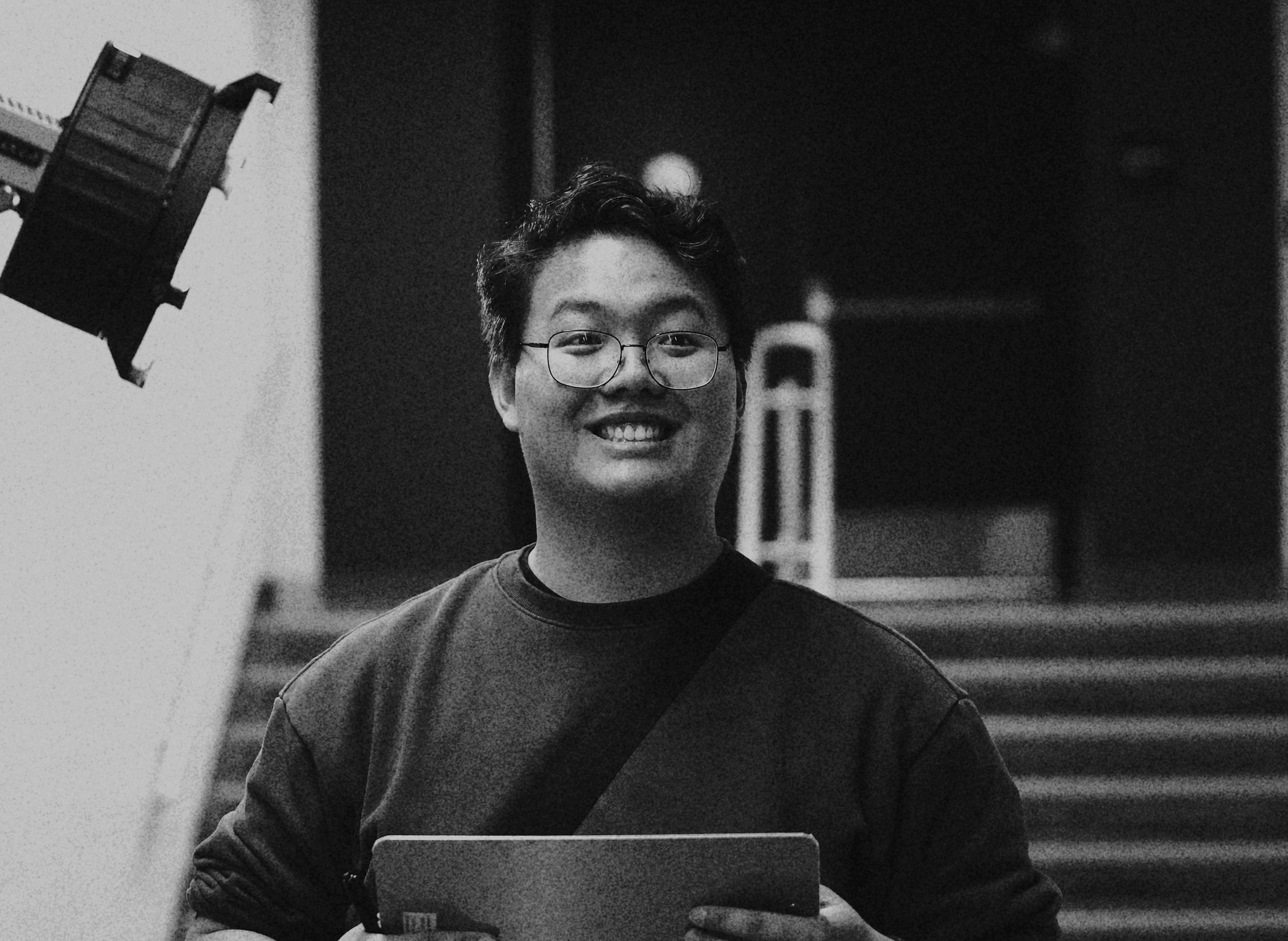 A person with glasses smiling in a black and white photo, holding a tablet in front of them, with studio lighting equipment and a background with various objects and textures.