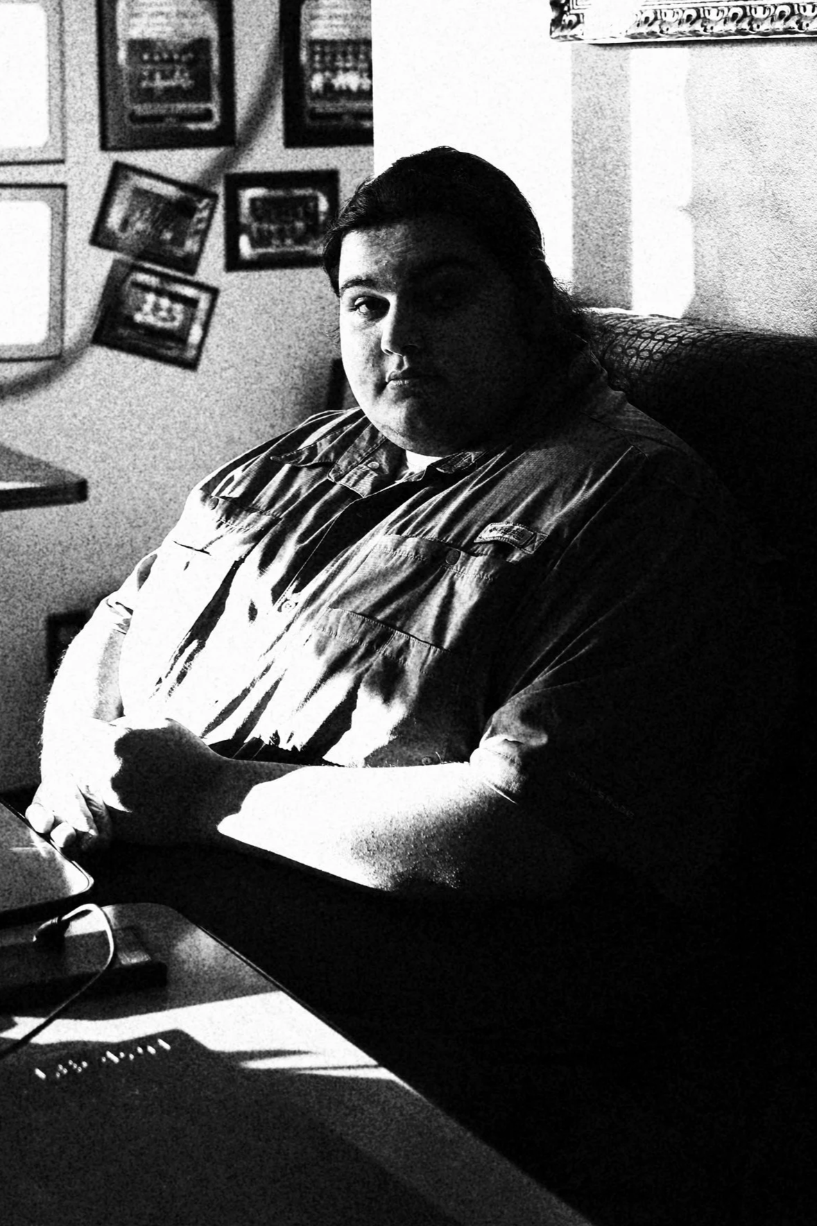 Black and white photo of a person with dark hair, sitting at a desk, with framed pictures on the wall behind them, in a room with some natural light streaming in.