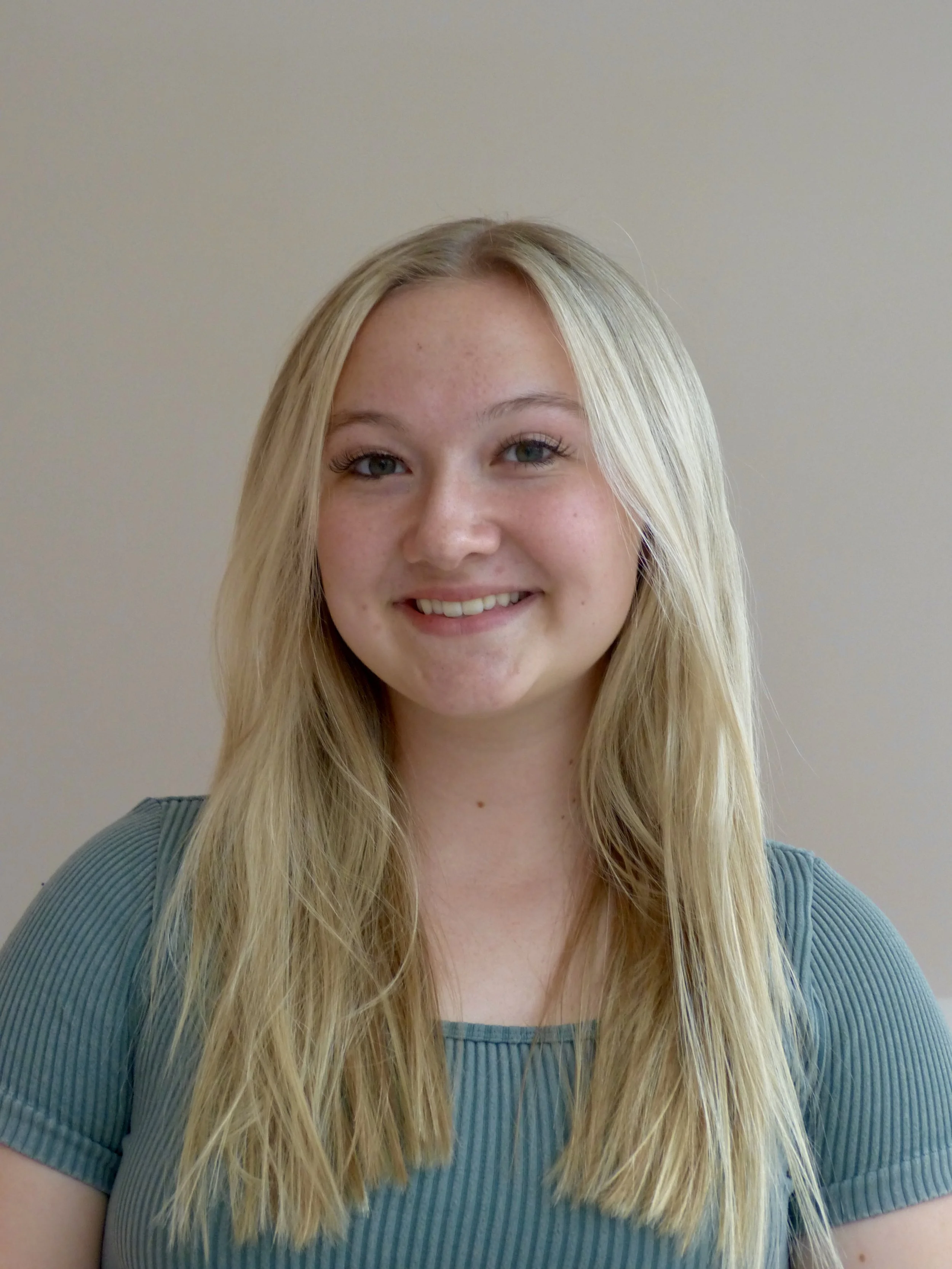 A young woman with long blonde hair, blue eyes, and a broad smile, wearing a teal ribbed shirt, standing against a neutral-colored wall.