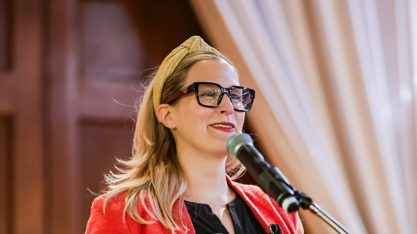 A woman with blonde hair, wearing glasses, a yellow headband, and a red blazer, speaking into a microphone in a room with wood-paneled walls and cream curtains.