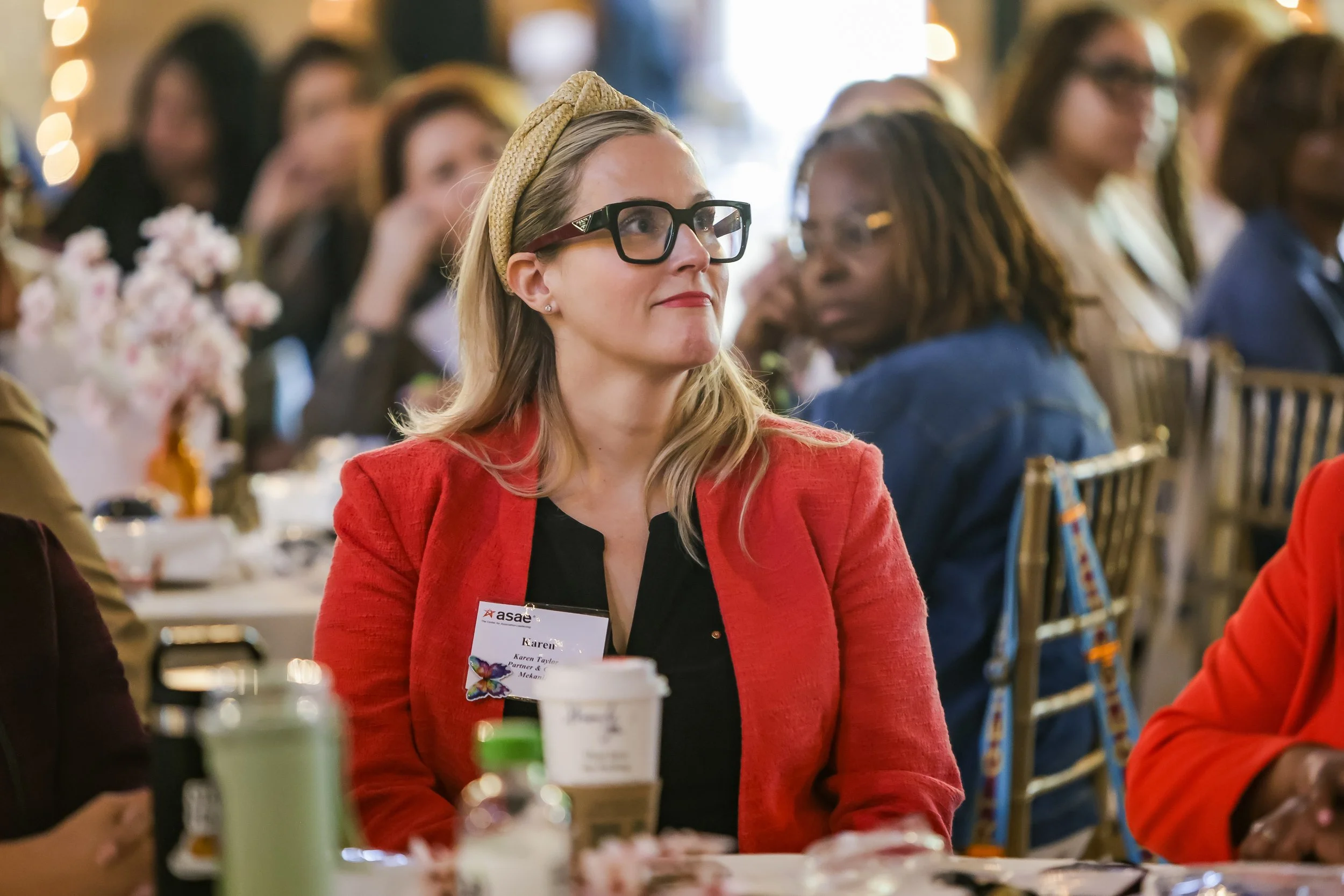 A woman with blonde hair wearing glasses, a beige headband, and a red blazer attending a conference or seminar. She has a name tag that reads 'Karen'. She is sitting at a table with various bottles and cups in front of her, and there are other people seated around her, blurred in the background.