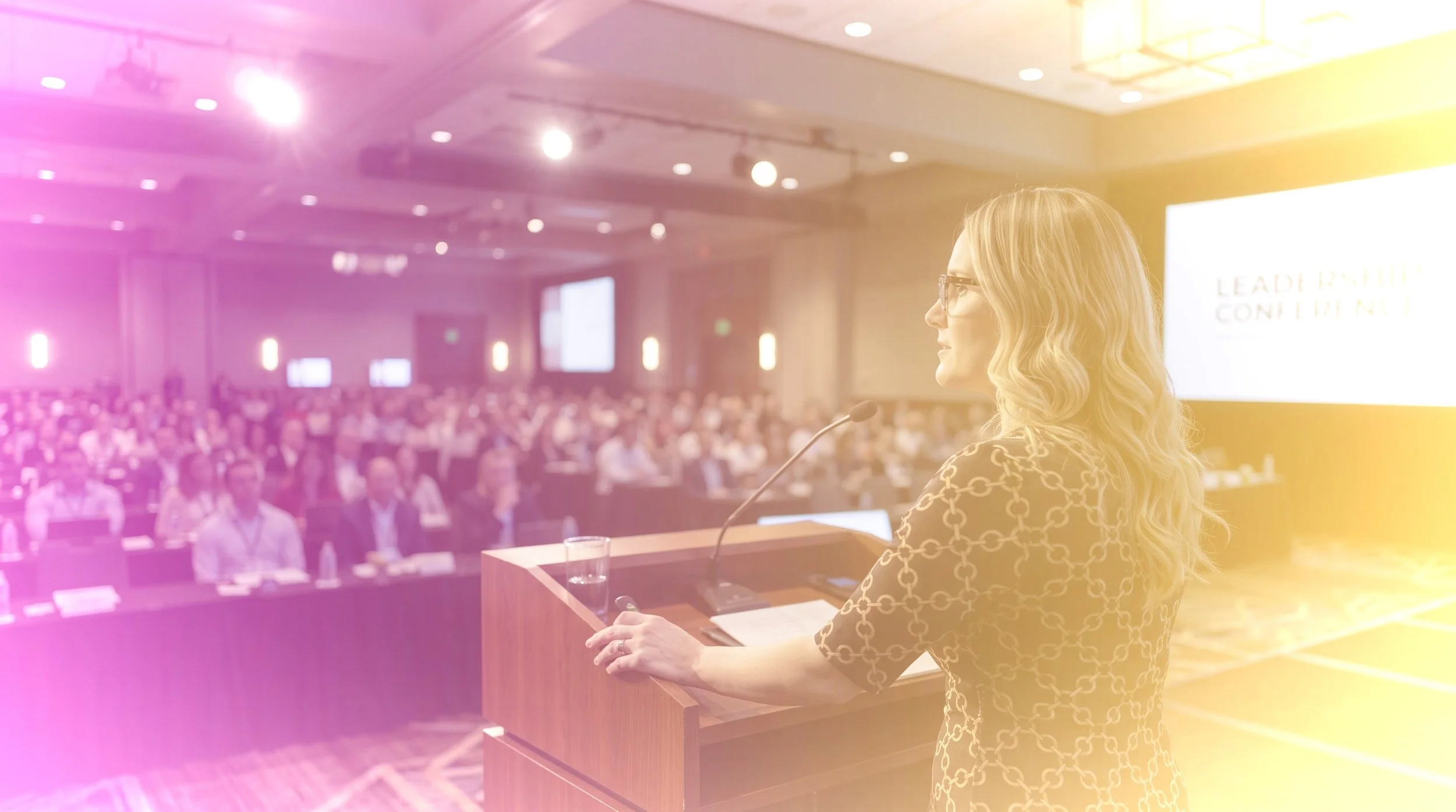 A woman with blonde wavy hair and glasses speaking at a conference, standing at a podium with a microphone, in a large conference room filled with seated attendees and a bright screen in the background.