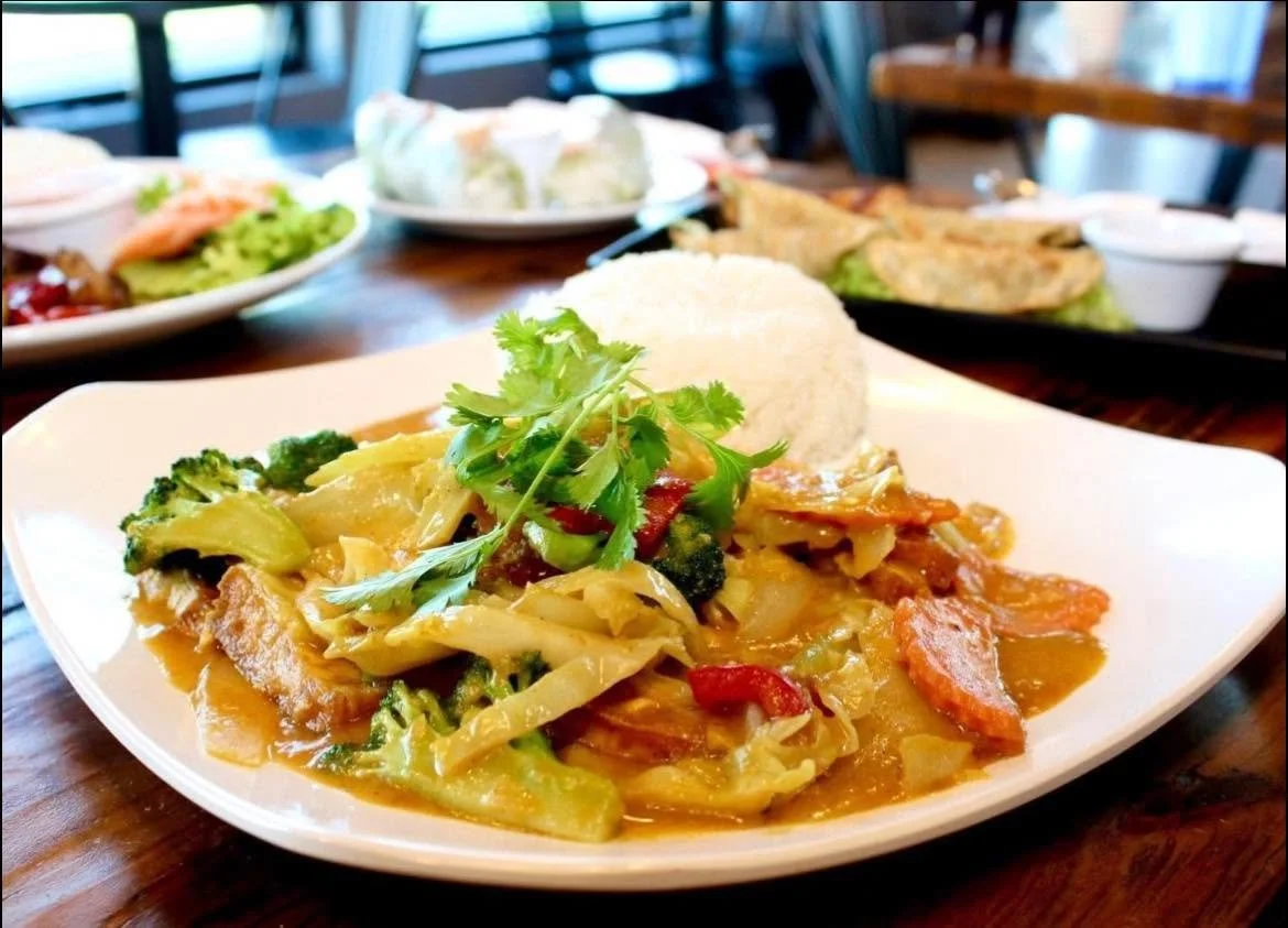 A plate with stir-fried vegetables, rice, and chicken garnished with cilantro, served on a white dish on a wooden table, with other plates of food in the background.