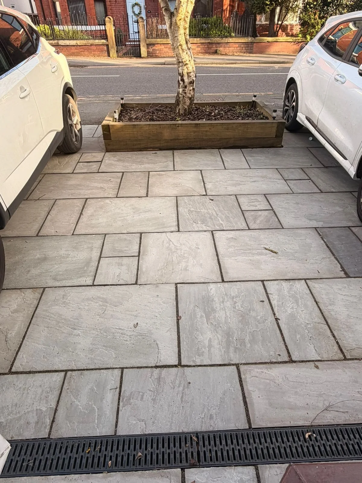 A paved sidewalk with large, rectangular stone tiles in front of two parked white cars. In the middle, there is a raised wooden planter box with a tree planted in it. A street with houses and cars is visible beyond the sidewalk.