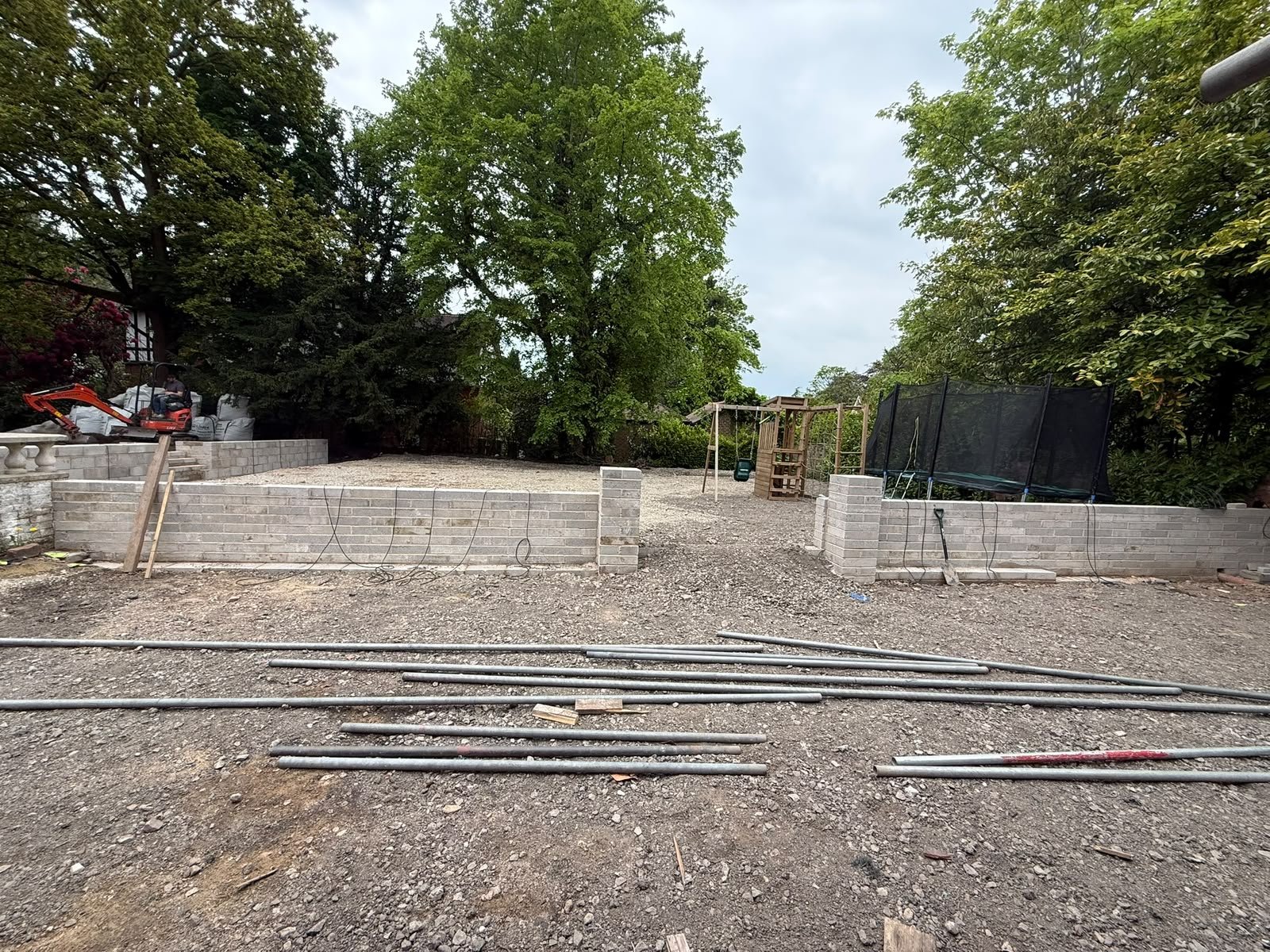 A backyard under construction with a partially built brick wall, construction materials, a small excavator, and playset equipment amid trees and a cloudy sky.
