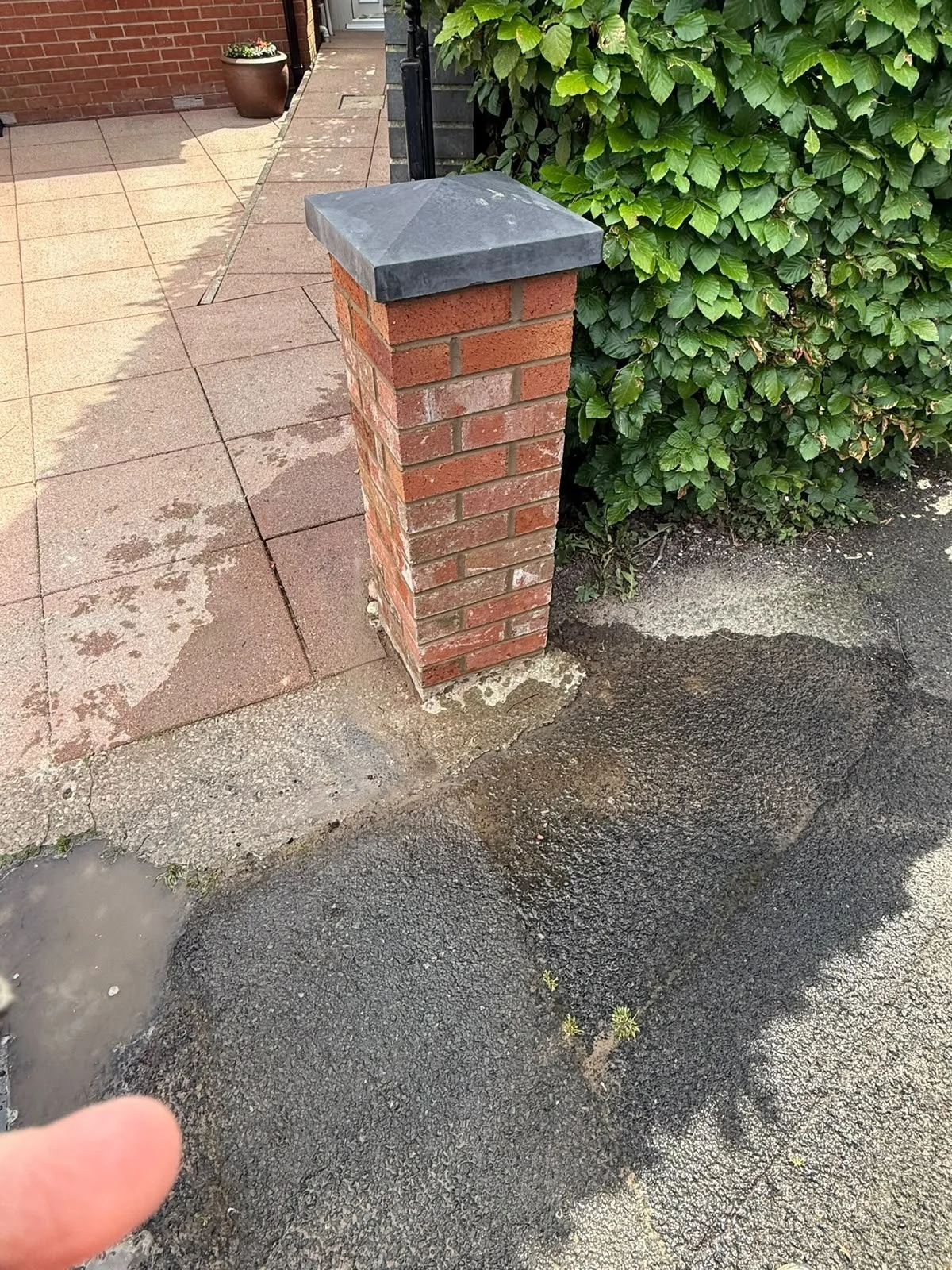Close-up of a brick pillar with a flat dark gray capstone at the edge of a paved area, next to a lush green bush, with wet pavement and a partial view of a house entrance in the background.