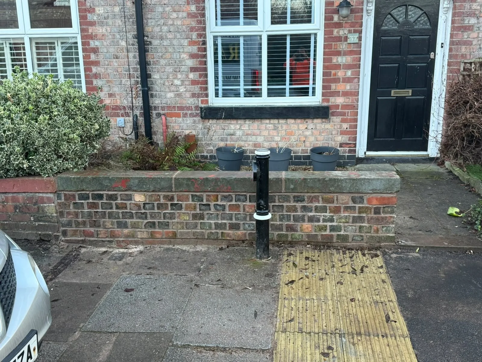 Front of a brick house with a black door, a window with white framing, three flower pots on a low brick wall, and a black post in the foreground, with a sidewalk and part of a parked car visible.