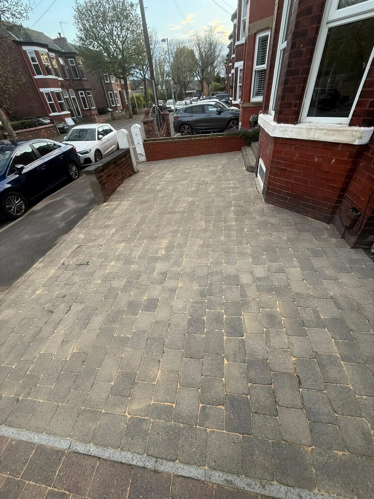 View of a residential front yard paved with brick pavers, adjacent to sidewalk with parked cars and brick houses in the background.