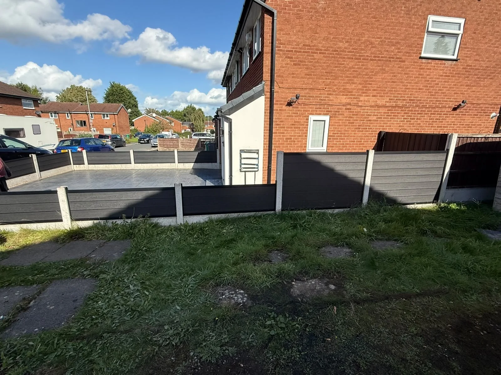View of a backyard with a small lawn and stepping stones leading to a fenced outdoor patio area. In the background, there are neighboring red brick houses and a parking lot with several cars. The sky is partly cloudy, and the scene is well-lit with s