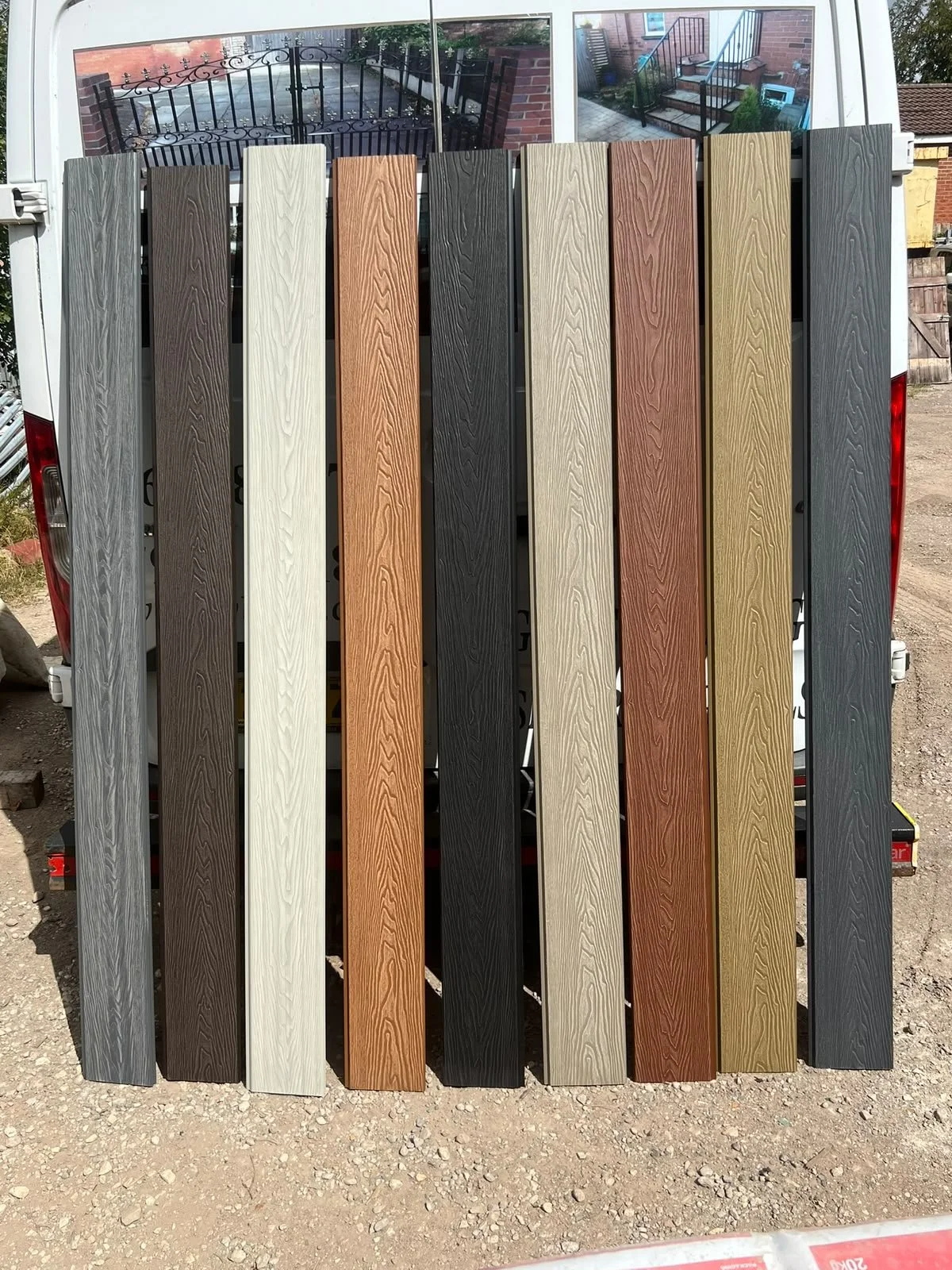A variety of outdoor decking planks in different colors leaning against a white trailer. The colors include shades of gray, brown, black, cream, and tan.