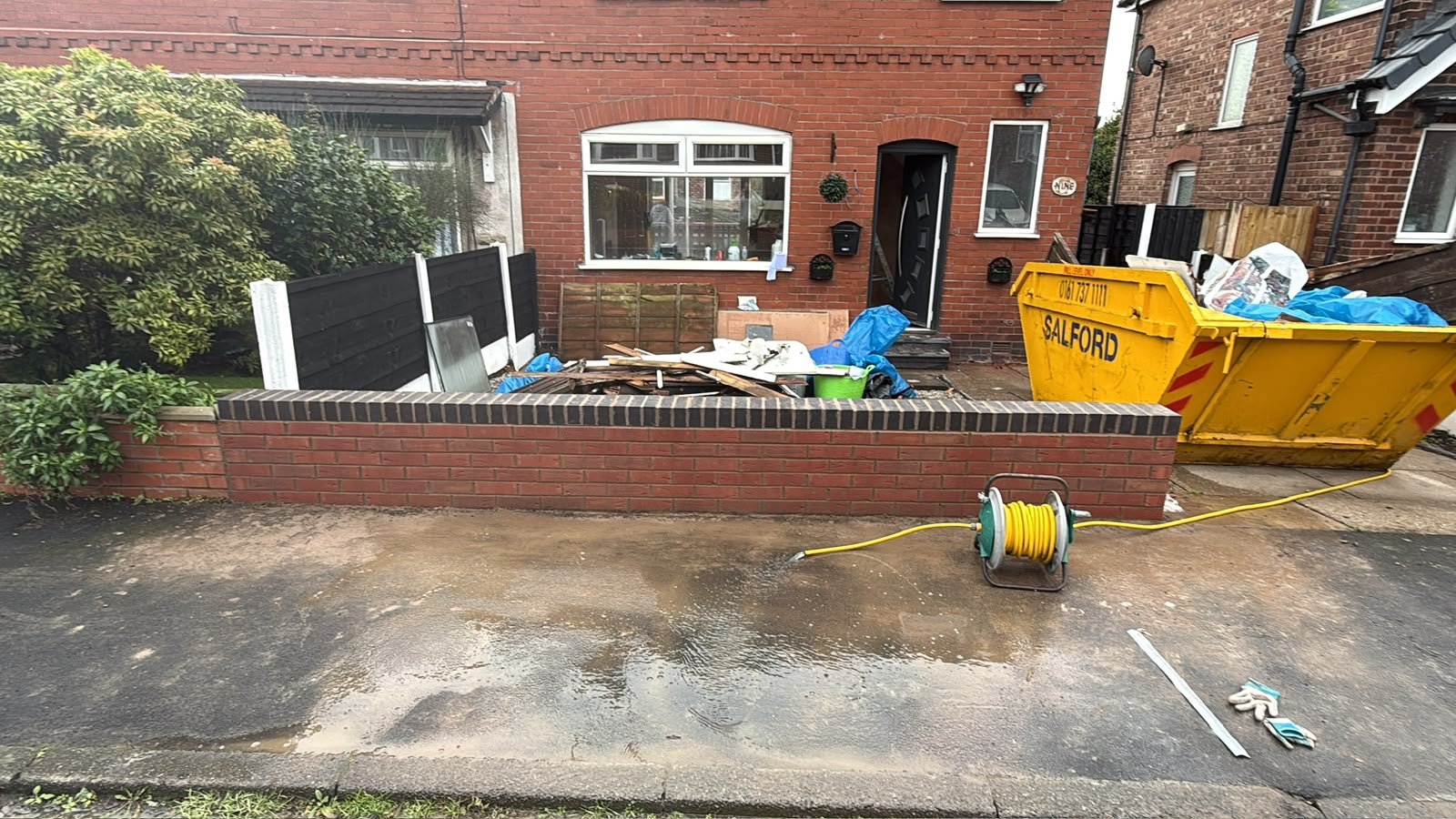A front yard of a brick house undergoing renovation, with construction tools, a yellow dumpster, a hose reel, construction debris, gloves, and plastic sheeting visible.