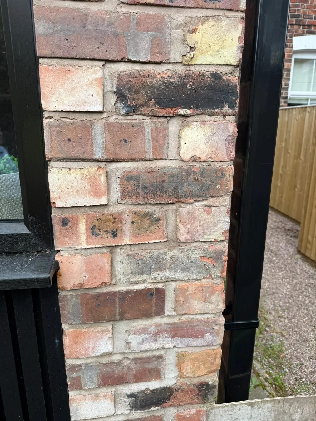Close-up of a brick wall with black metal framing on the sides, showing weathered bricks with varying shades of red, cream, and black, and some mortar stains.