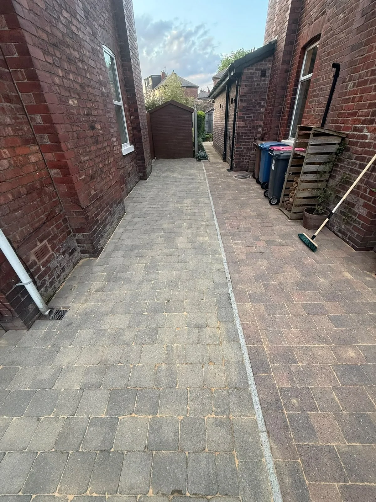 An alleyway between two brick houses with paving stones, a small shed at the end, and cleaning tools leaning against the wall.