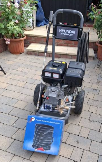 A pressure washer on a paved patio with a hose and a blue box in front containing a coil of extension cord or hose.
