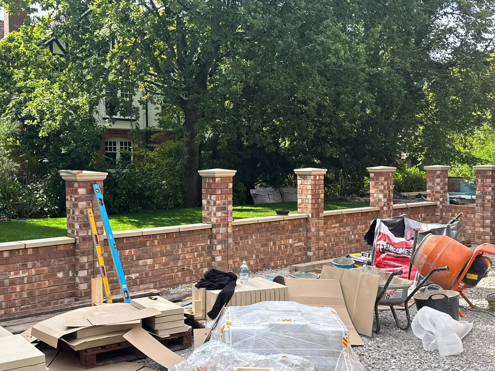 Construction site with a brick wall being built, construction tools and materials scattered around, including a wheelbarrow, ladder, and bags of supplies, in a backyard with large trees and a house in the background.