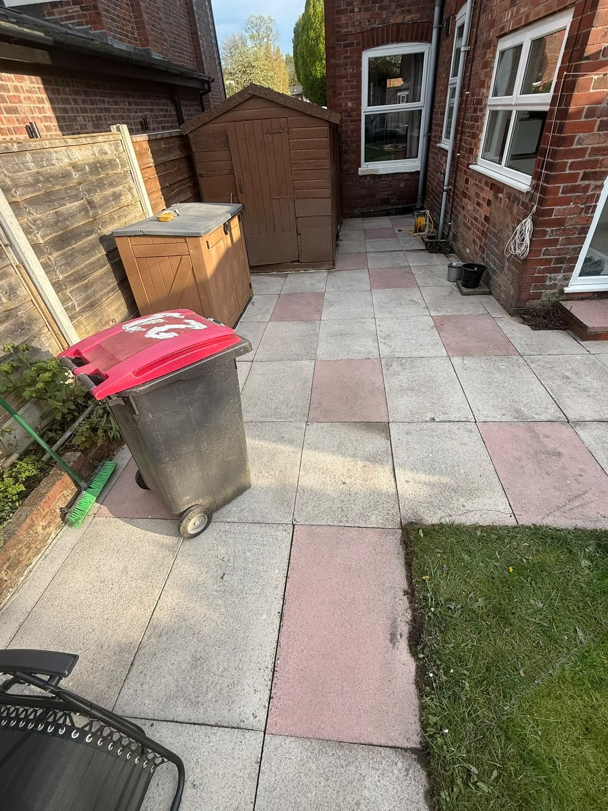 A small backyard patio with large concrete tiles, a wheelie trash bin with a red lid in the foreground, a green broom, a wooden shed, a wooden storage box, and a small garden bed along a brick wall. There are windows on the brick house and some lush 