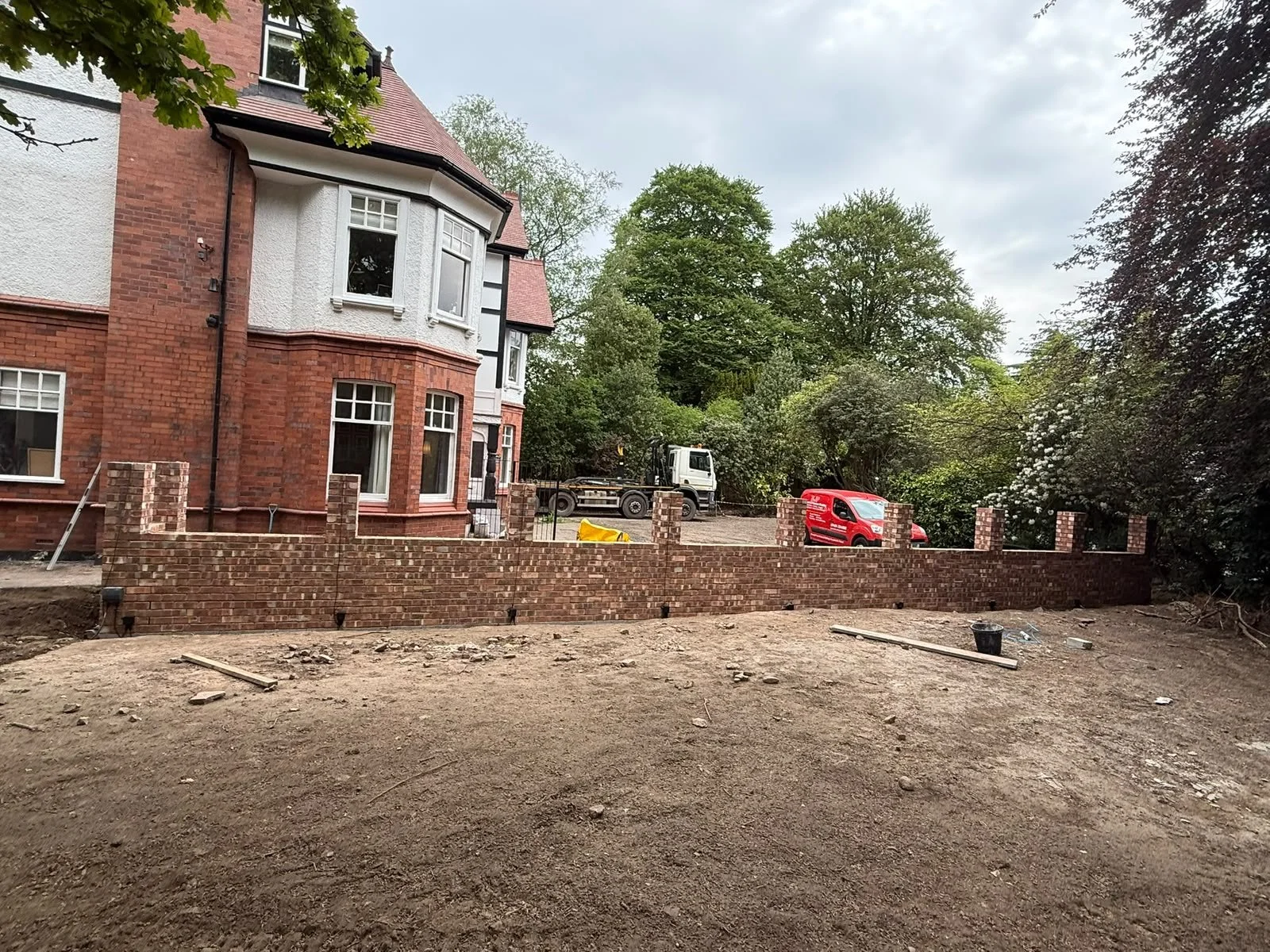A construction site in front of a red brick house with white window frames, with a brick wall partially built, construction tools and vehicles including a truck and a red van, and trees in the background.