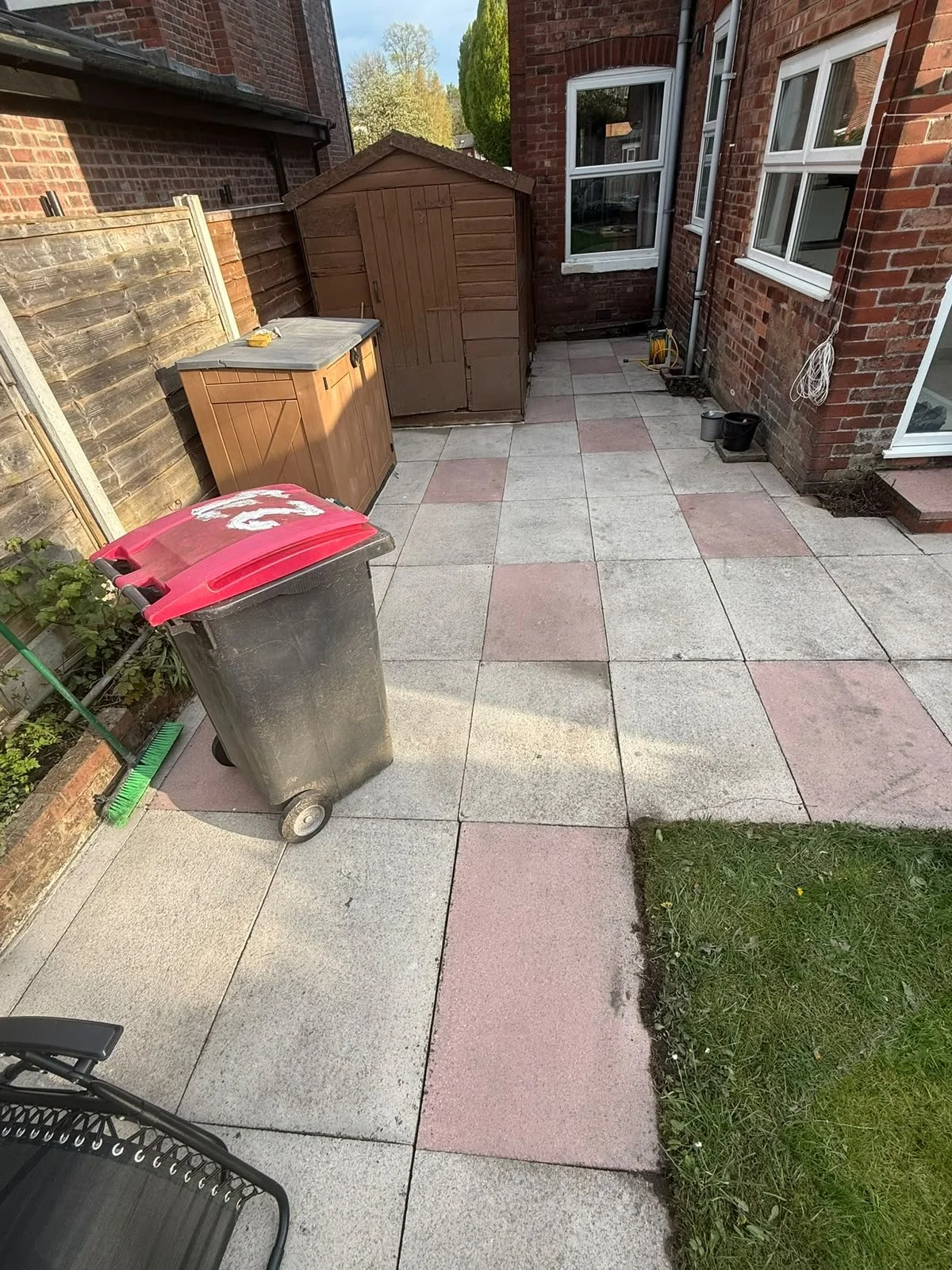 An outdoor patio area with tiled flooring next to a brick house. There is a trash bin with a red lid, a small wooden shed, a storage box, some gardening tools, and a few black buckets. Part of a green lawn is visible in the lower right corner.