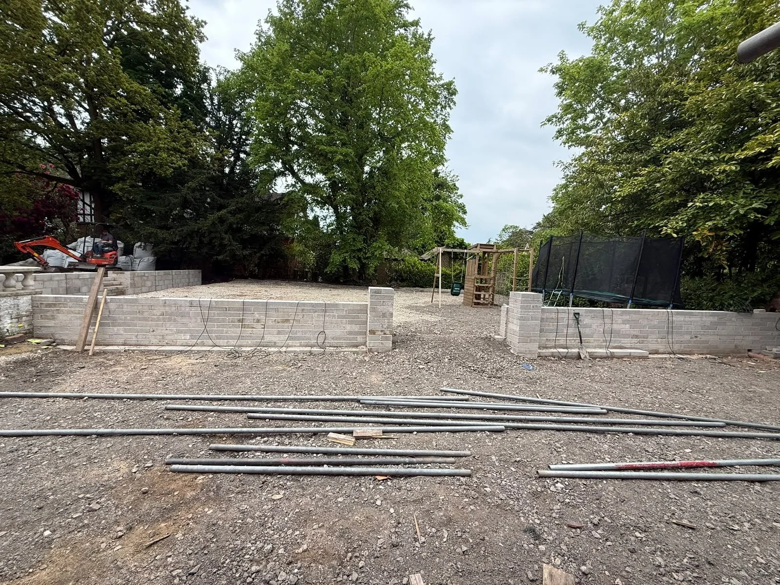 A backyard under construction with a partly built brick wall, construction materials, a small excavator, and a playground set with a swing and a trampoline, surrounded by trees.