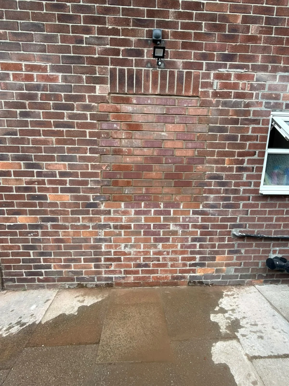 Backside of a brick building with a small window, a security camera, a floodlight, and a pipe. The brick wall has a section with different bricks. The ground shows wet patches and concrete slabs.