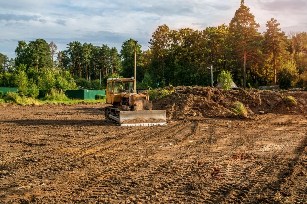 A construction site with a yellow bulldozer on bare, disturbed earth, surrounded by dirt mounds and trees in the background during late afternoon.