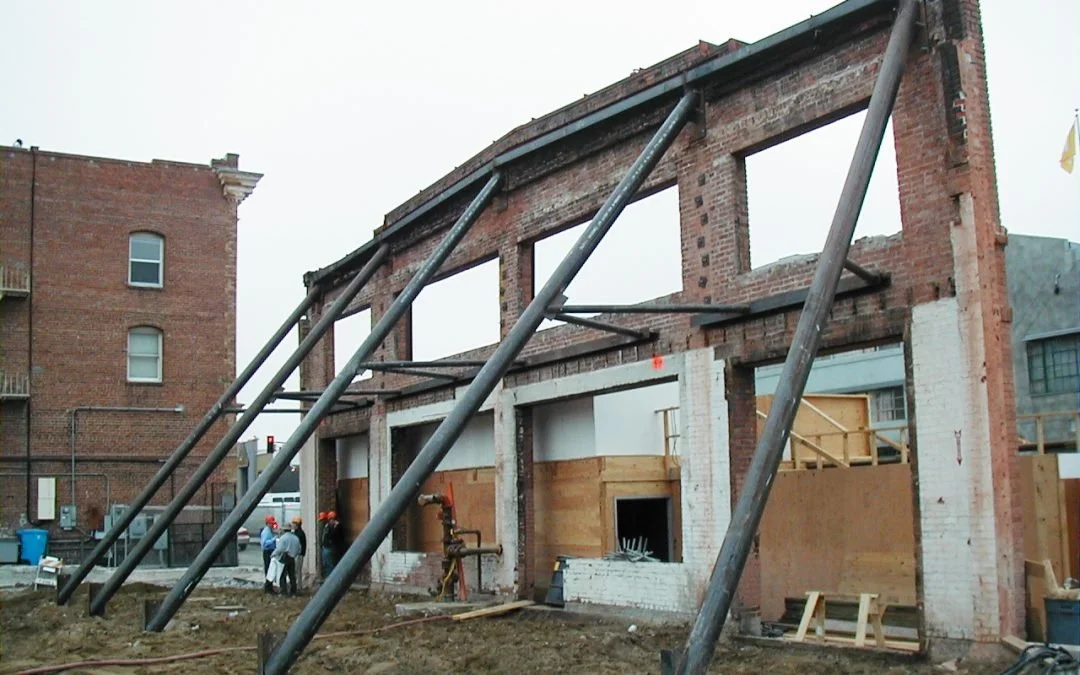 Under construction brick building with temporary support beams, scaffolding, and workers in the background.