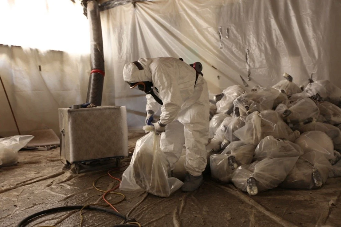 A person wearing a white hazmat suit, gloves, and mask working with plastic bags filled with material in a makeshift environment.
