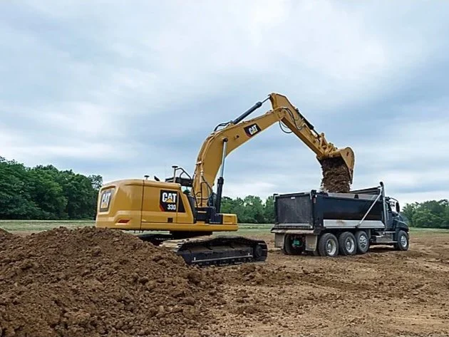 A yellow CAT excavator loading dirt into a black dump truck on a construction site with trees in the background under a cloudy sky.