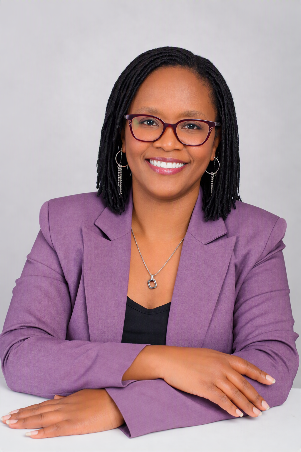 A professional woman with glasses and earrings, wearing a purple blazer and a necklace, smiling at the camera with arms crossed.