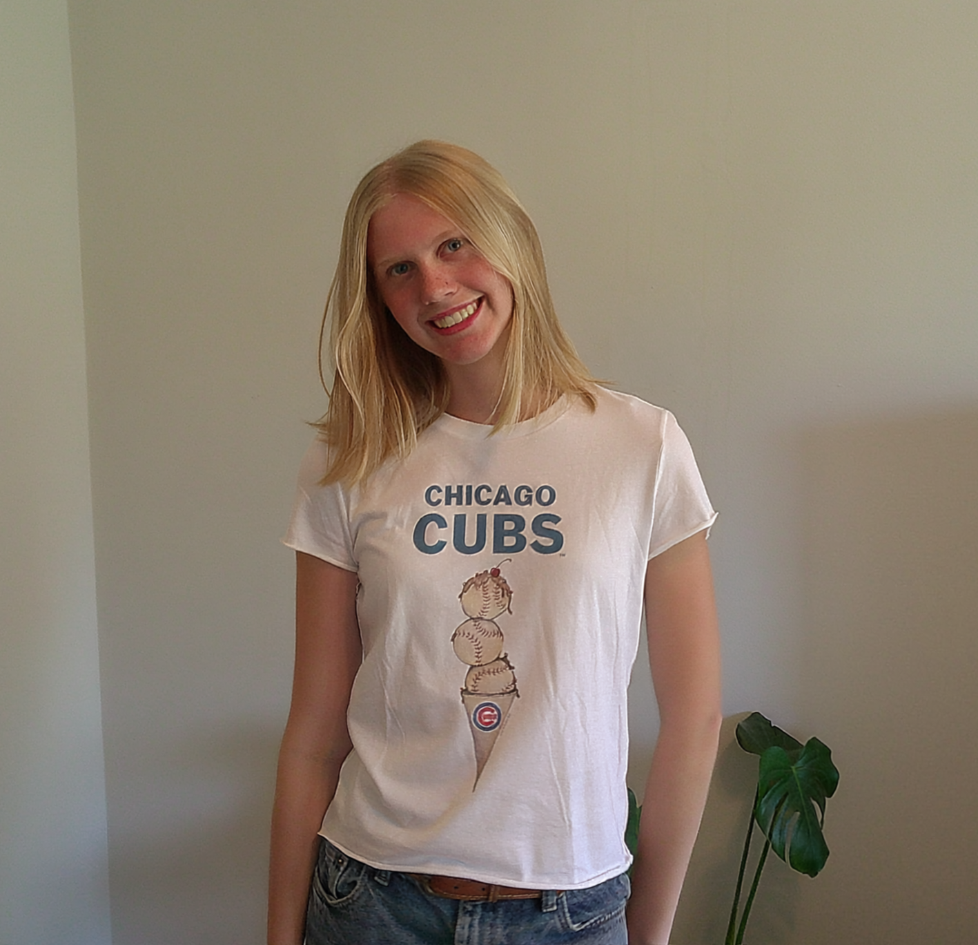 A woman with blonde hair wearing a White Chicago Cubs T-shirt and jeans standing near a potted plant indoors.