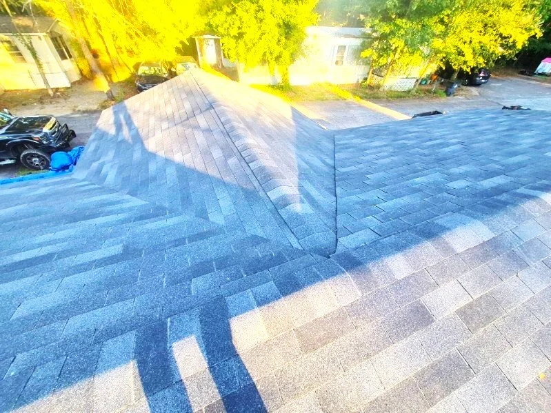 View of a residential roof with asphalt shingles, showing multiple slopes and a chimney, with trees and houses in the background.