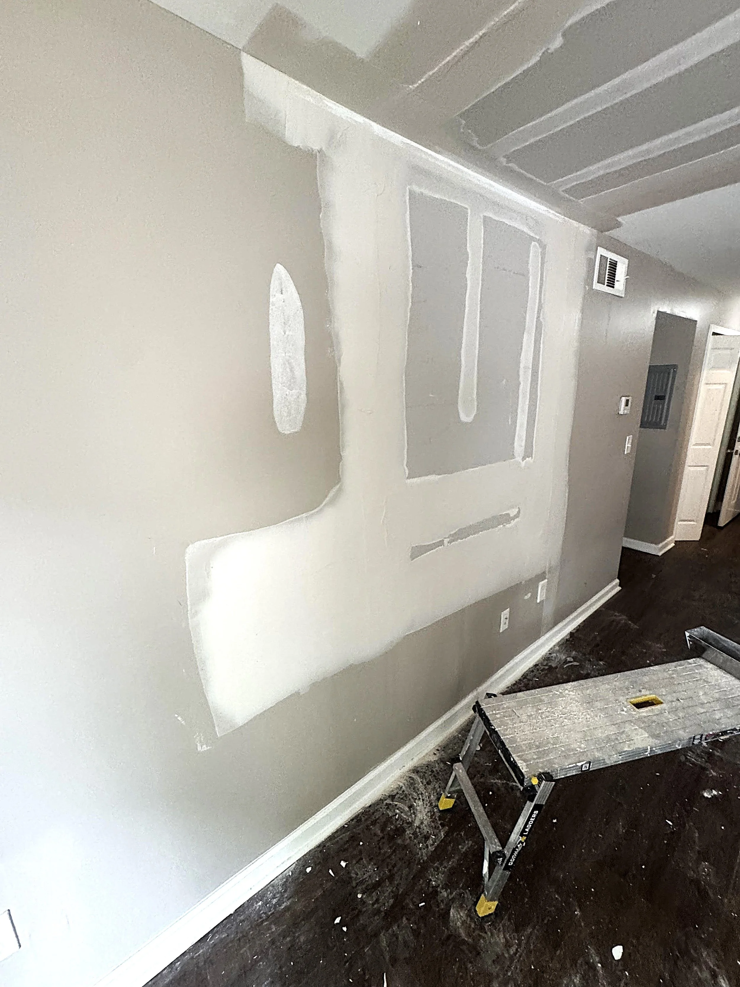 A partially finished interior wall with drywall and joint compound, a step ladder on a dark hardwood floor, and a thermostat on the wall in a home under renovation.